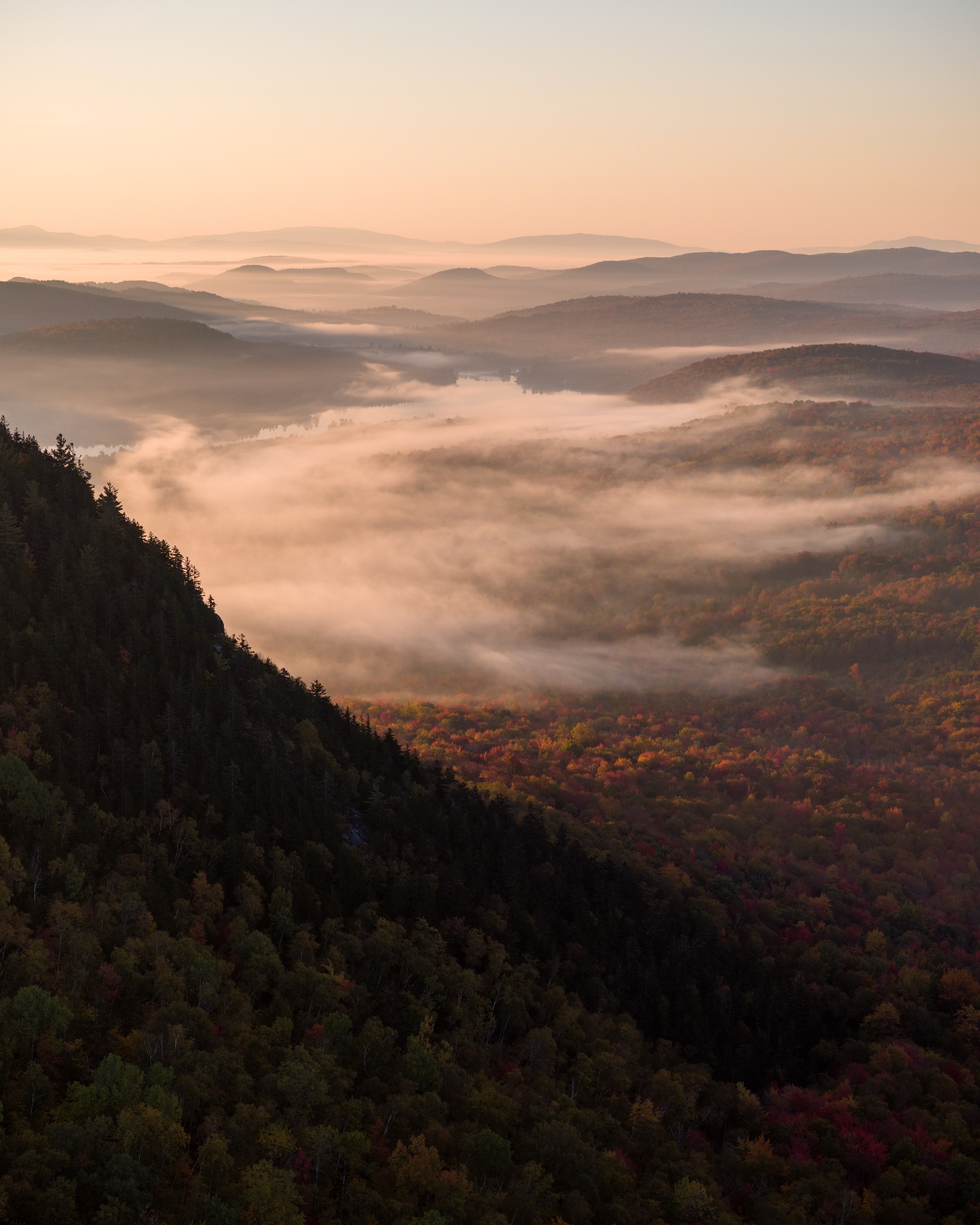 a photo of a valley and mountainscape of rolling vermont hills, covered in a mush of red fall foliage and early morning fog.
